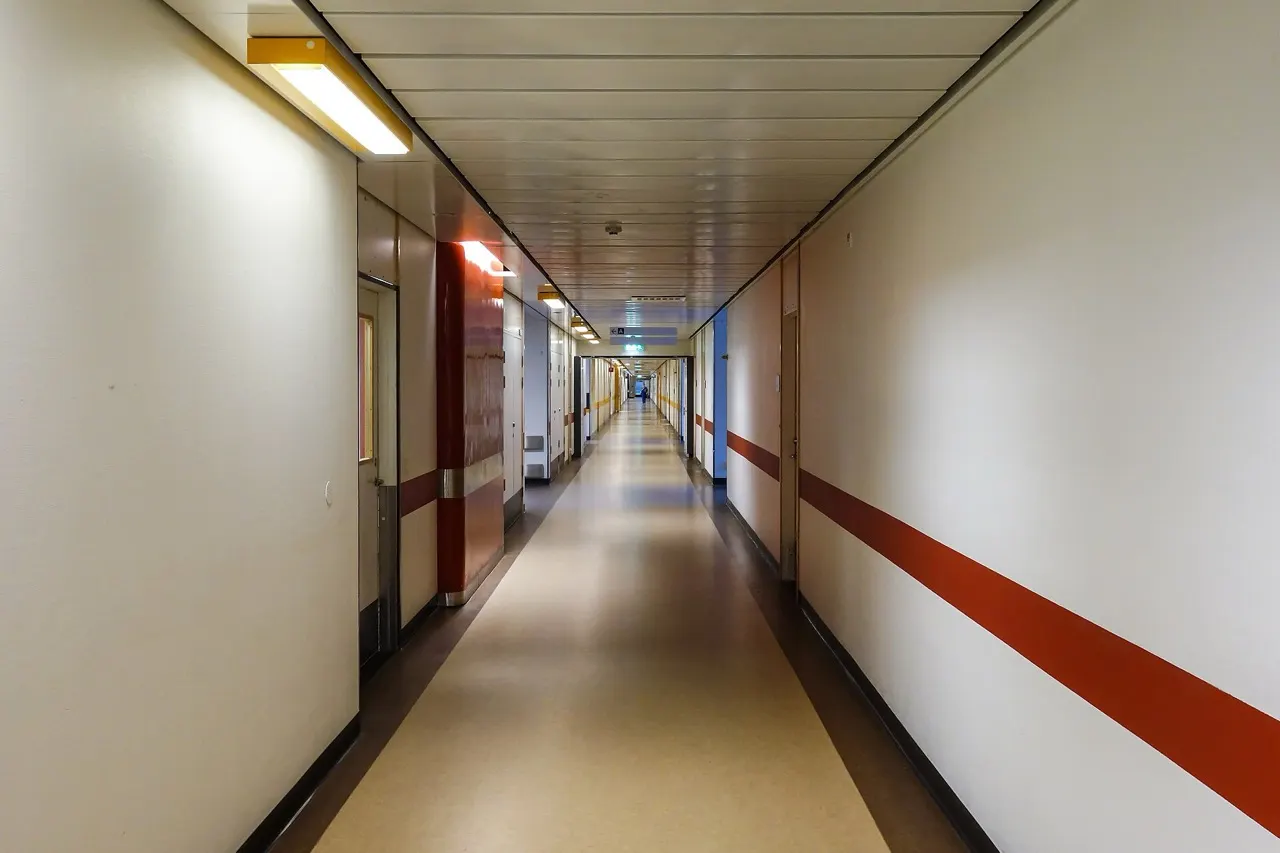 A long, empty hospital corridor with cream walls, a red stripe, and ceiling lights