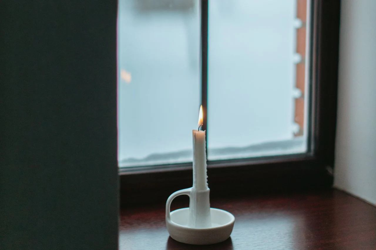 A single lit white candle in a ceramic holder on a dark wooden windowsill, with snow outside the window
