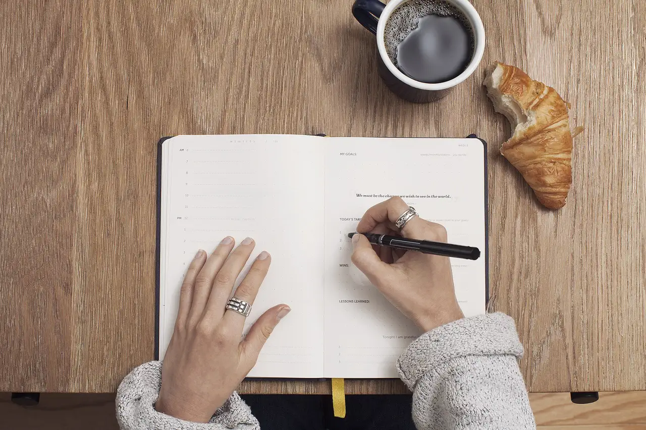 Hands writing in an open notebook beside a cup of coffee on a wooden table