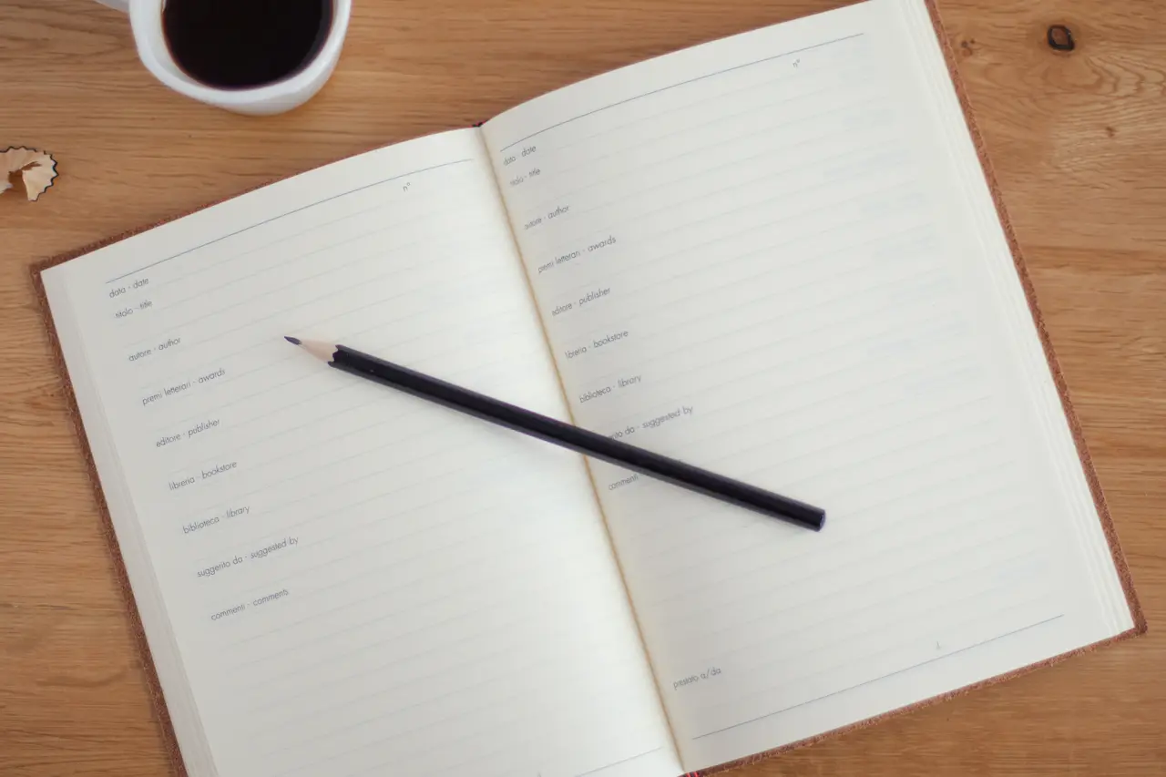 An open notebook with a pencil beside a cup of black coffee on a wooden table